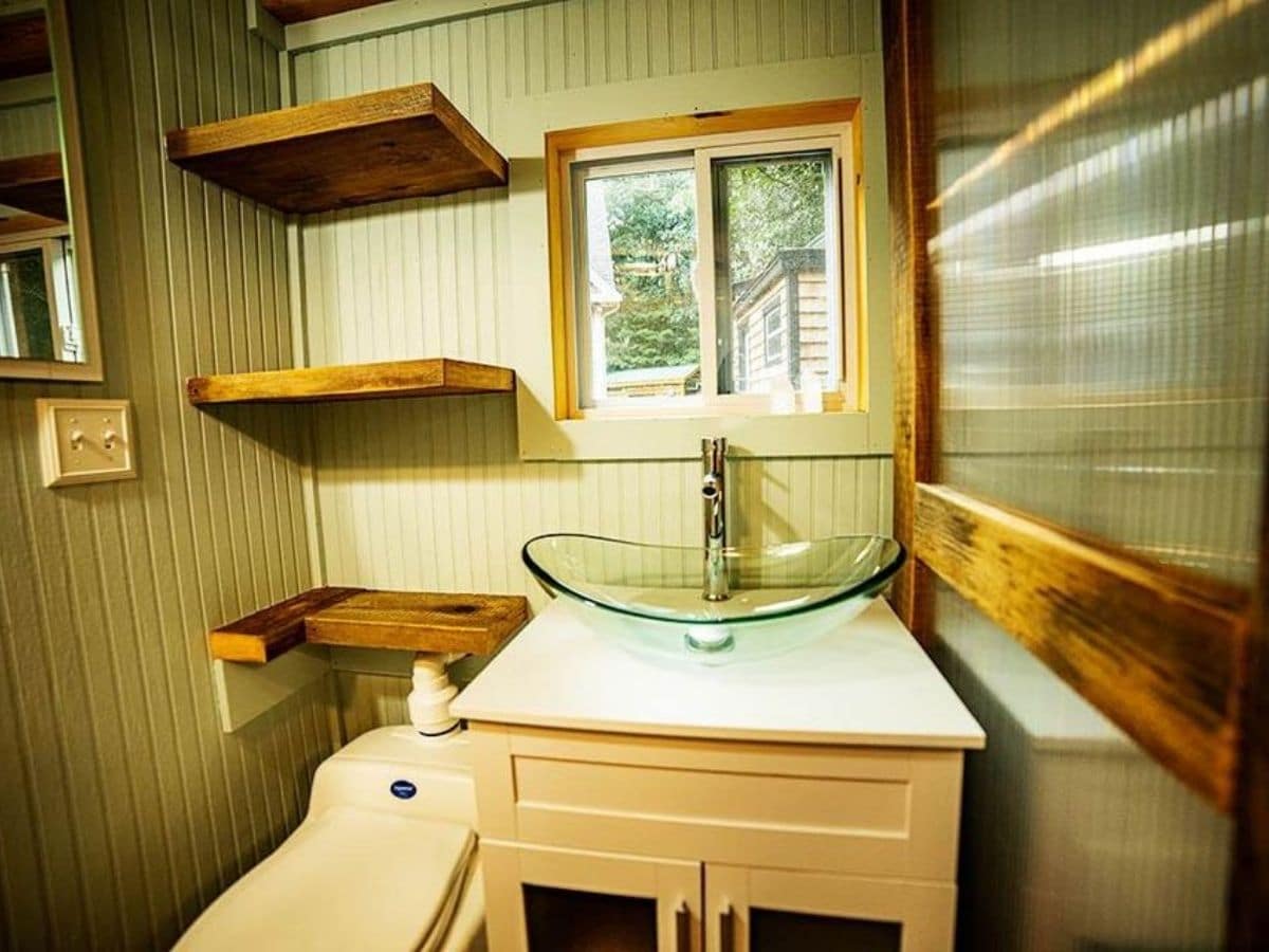 Glass sink above white vanity by toilet with wood shelves above