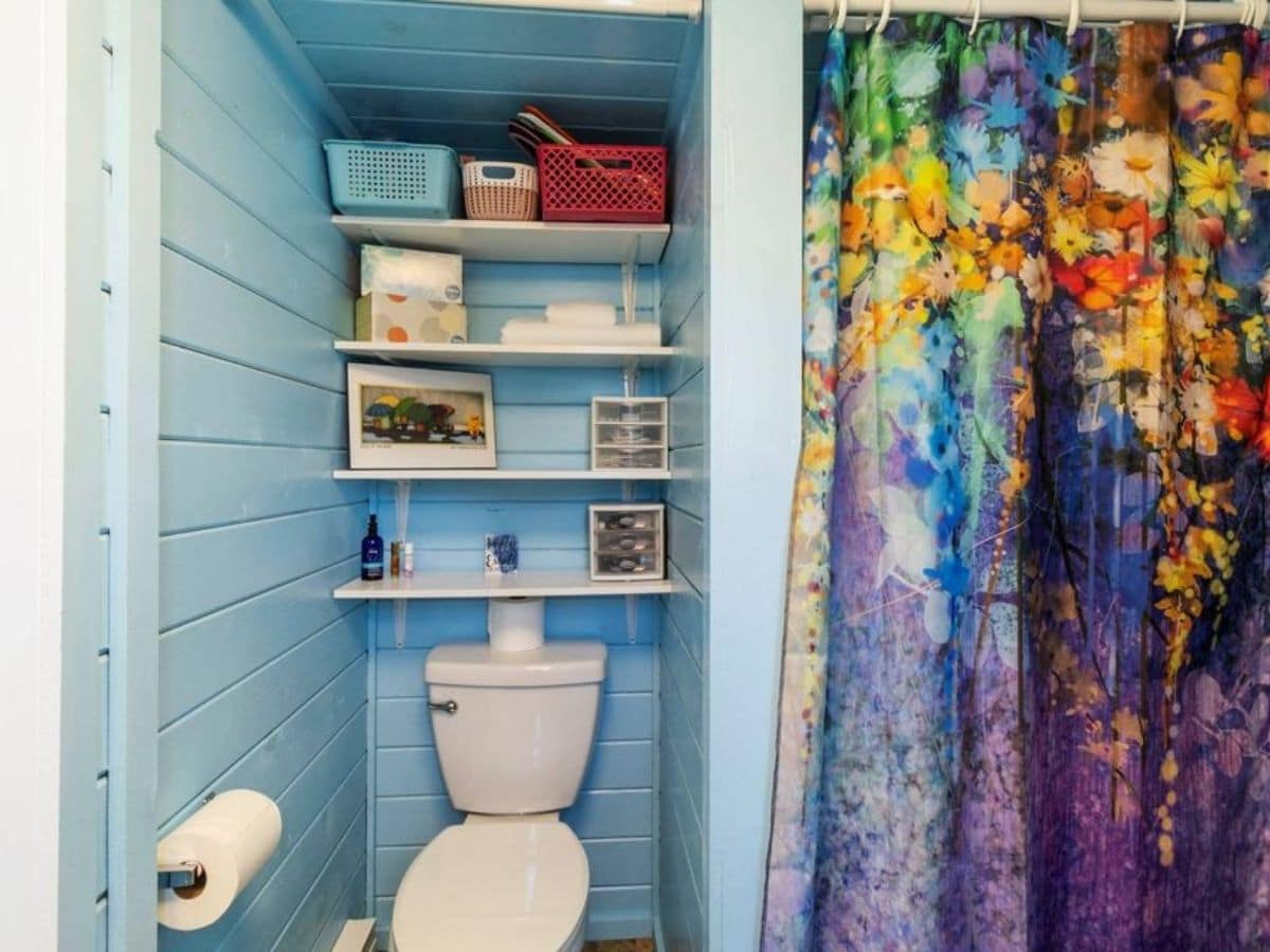 teal walls in bathroom with white flush toilet below open shelves and colorful shower curtain on right