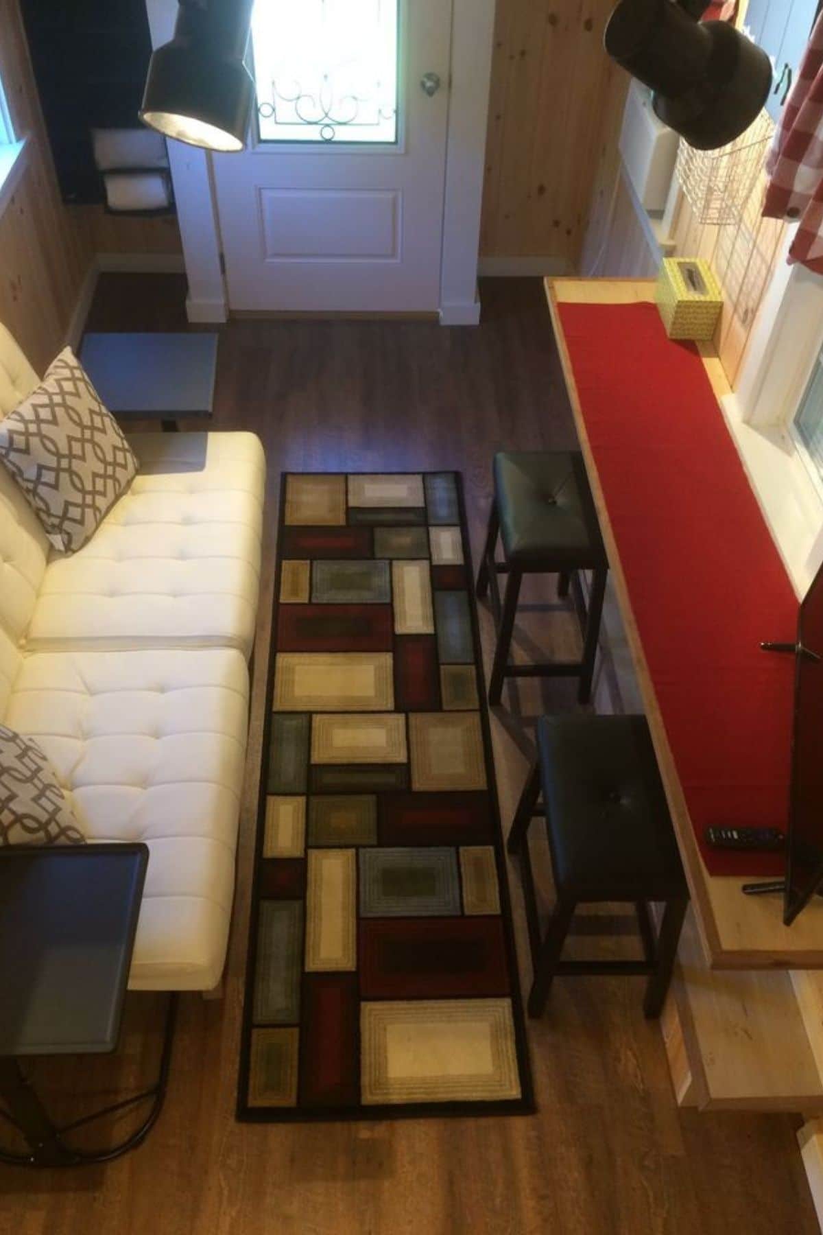view down on living room from tiny home loft showing white sofa, counter under window, and colorful rug in center of room
