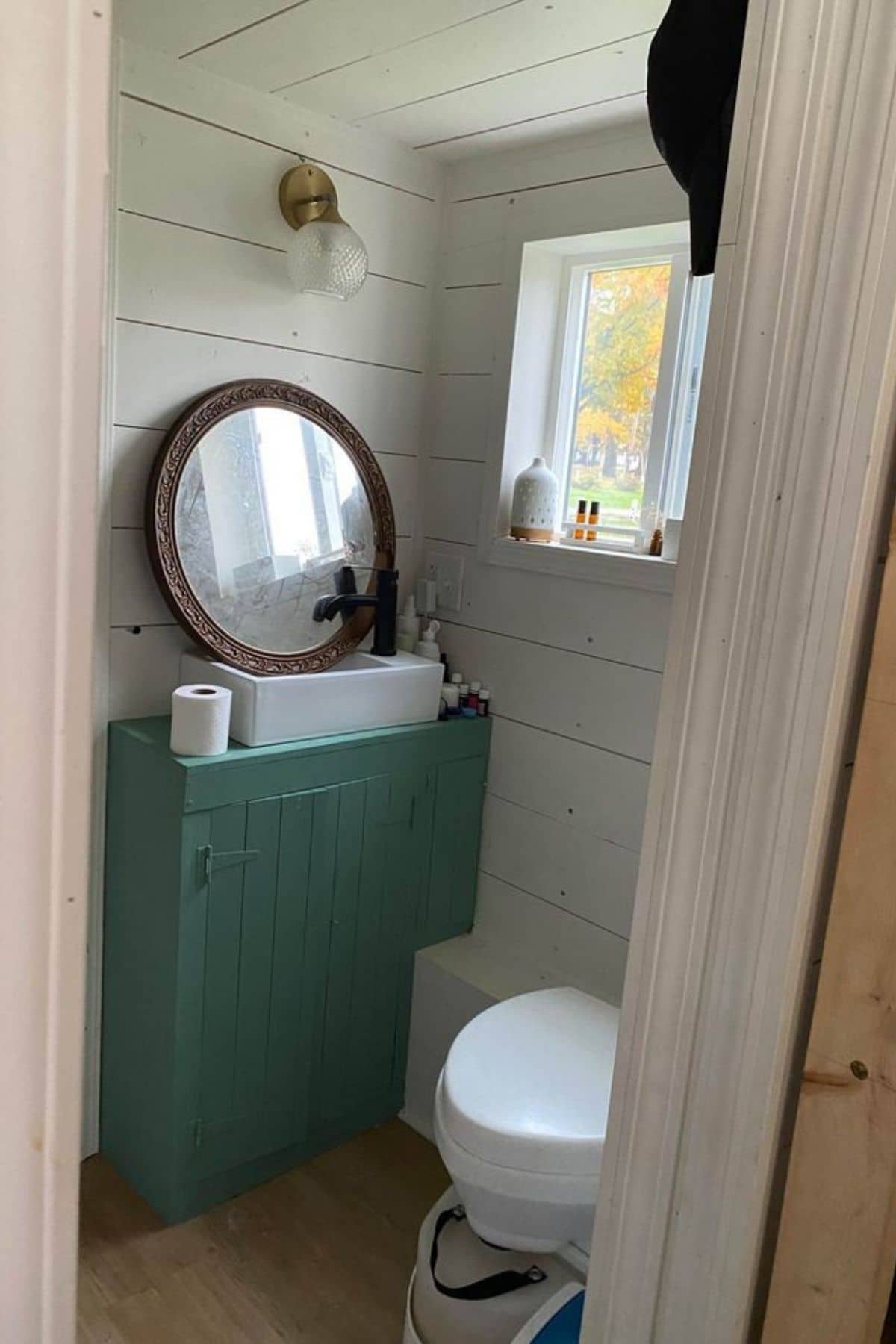teal vanity under round mirror next to white toilet under window