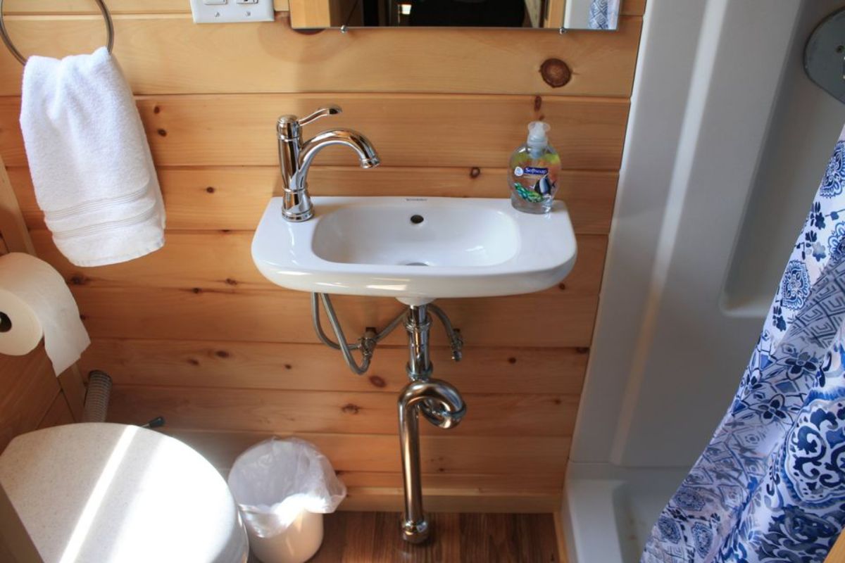 white sink against wall with open pipes and shower on right with toilet on left