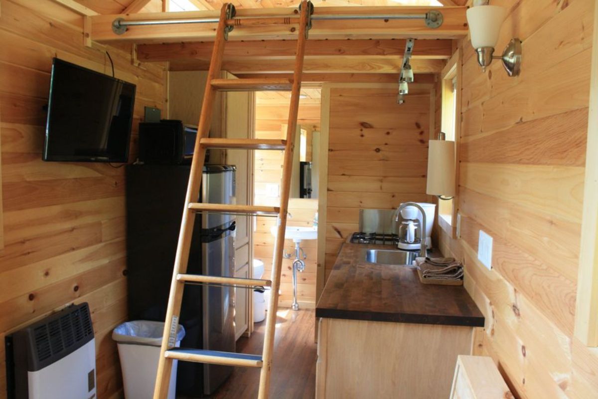 ladder leaning agaisnt loft in middle of tiny home with wood cabinet on right and refrigerator on left