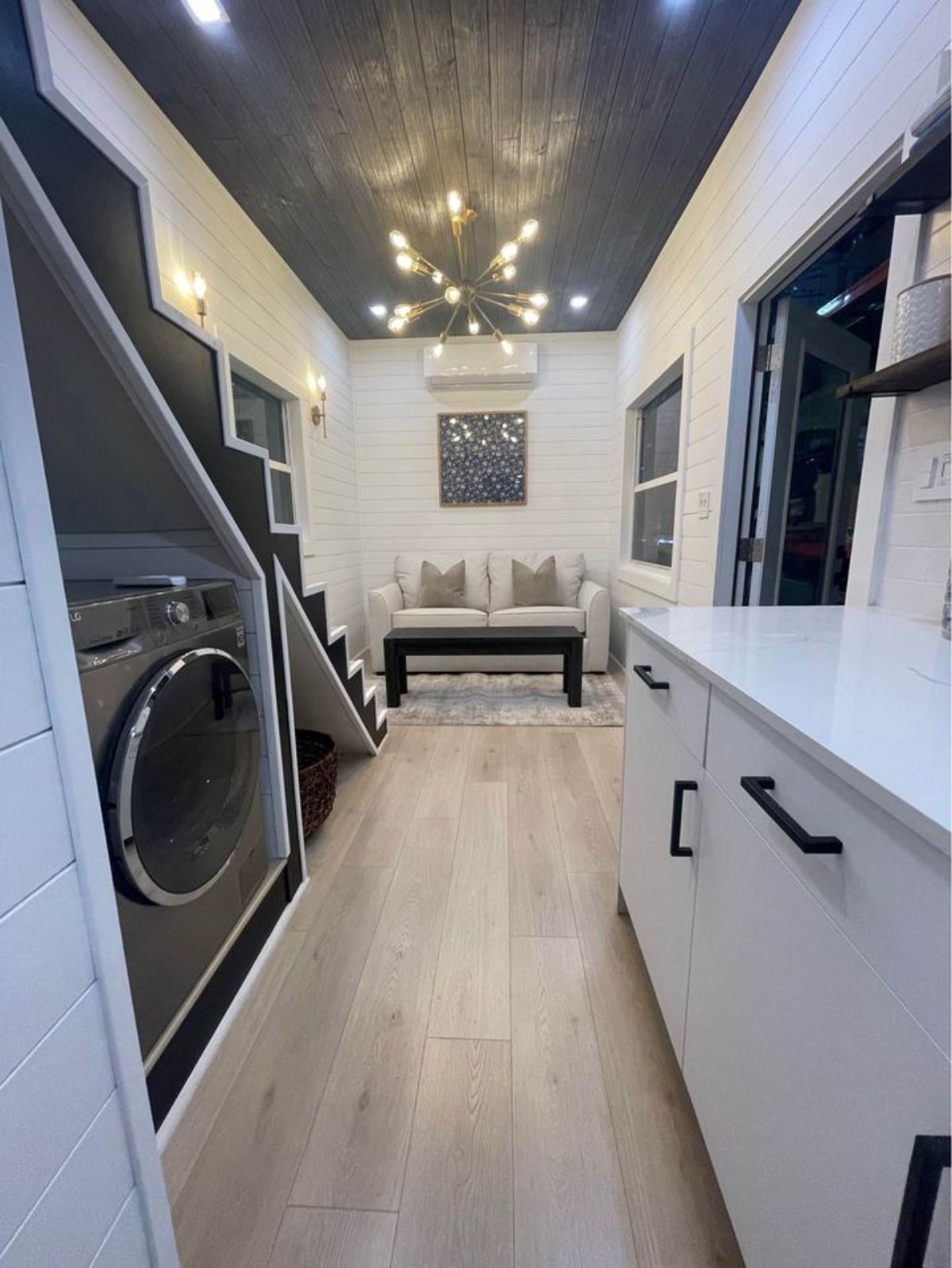 stainless steel combination washer and dryer on left under stairs with white cabinets and counter on right with black hardware