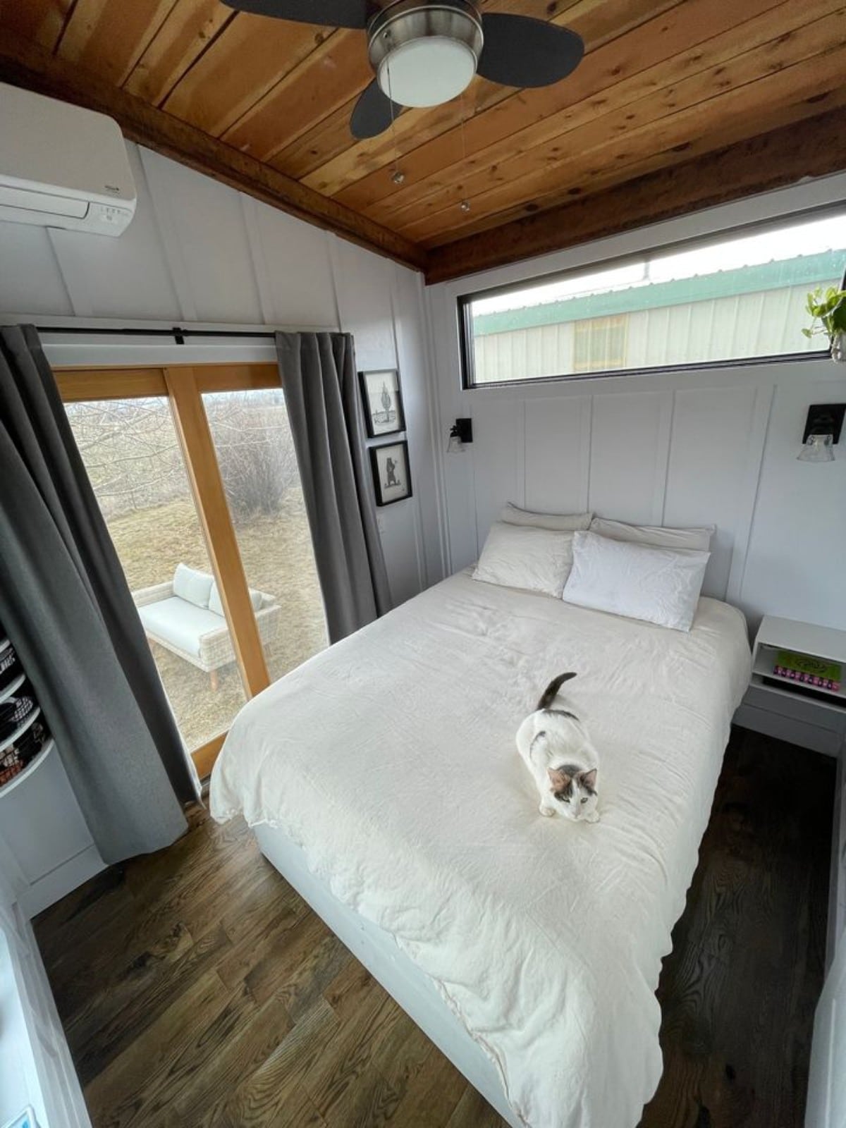 Bedroom of 30’ Tiny House on Wheels has classy custom made queen bed, a side table, shelves, an air condition unit with huge glass window and back door