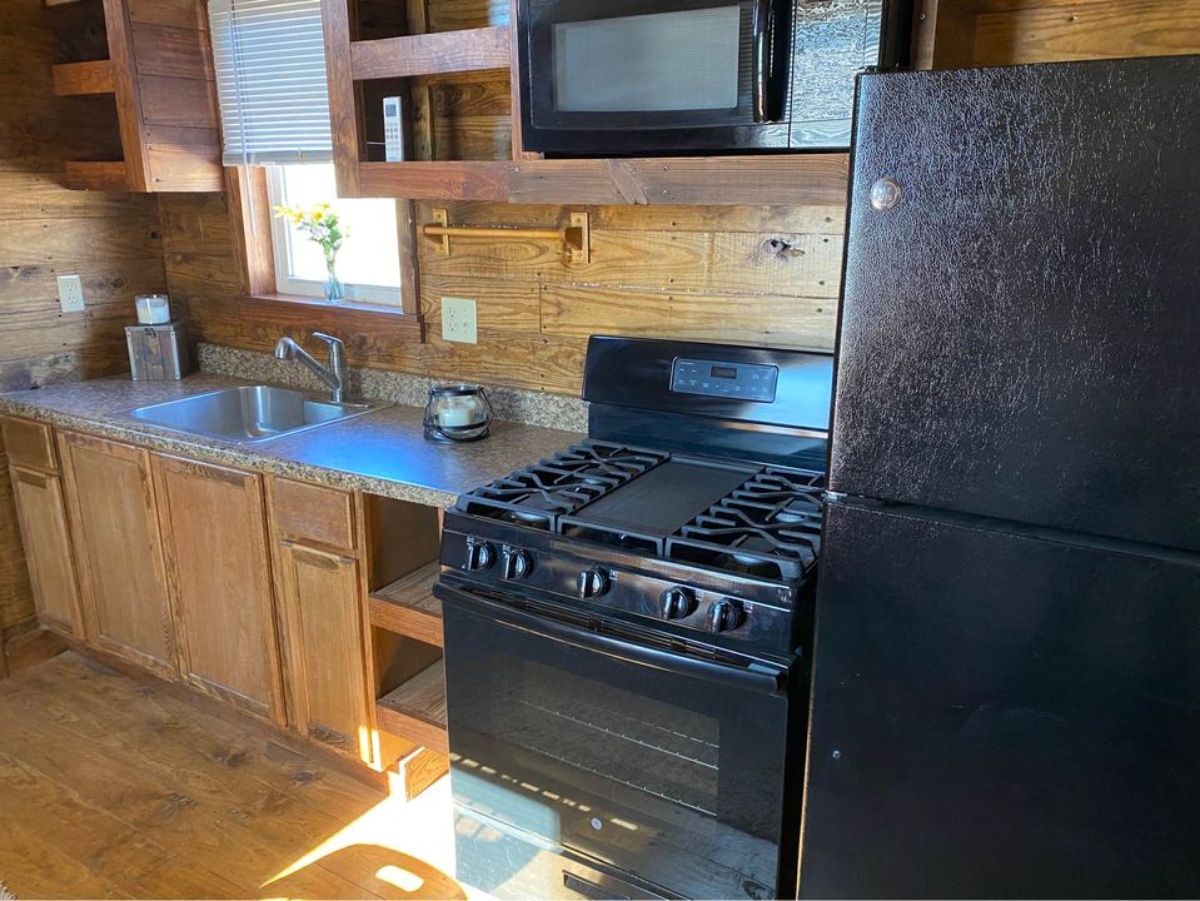Kitchen area of 36' Tiny House has 4 burner stove and oven, a full-sized refrigerator, a stainless steel sink, and a hanging microwave oven.