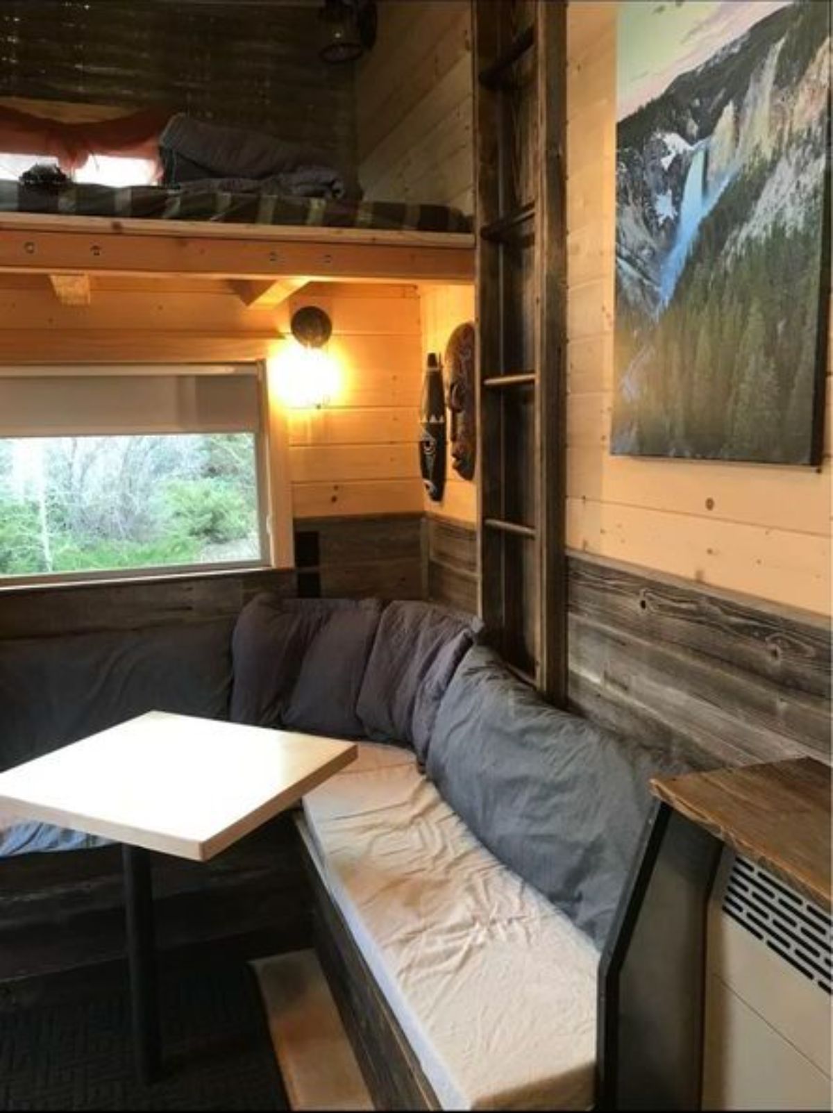 The living area of 24’ Rustic Tiny House On Wheels has an L shaped couch, removing table, wall mounted book shelf and a loft which can be used as extra sleeping space