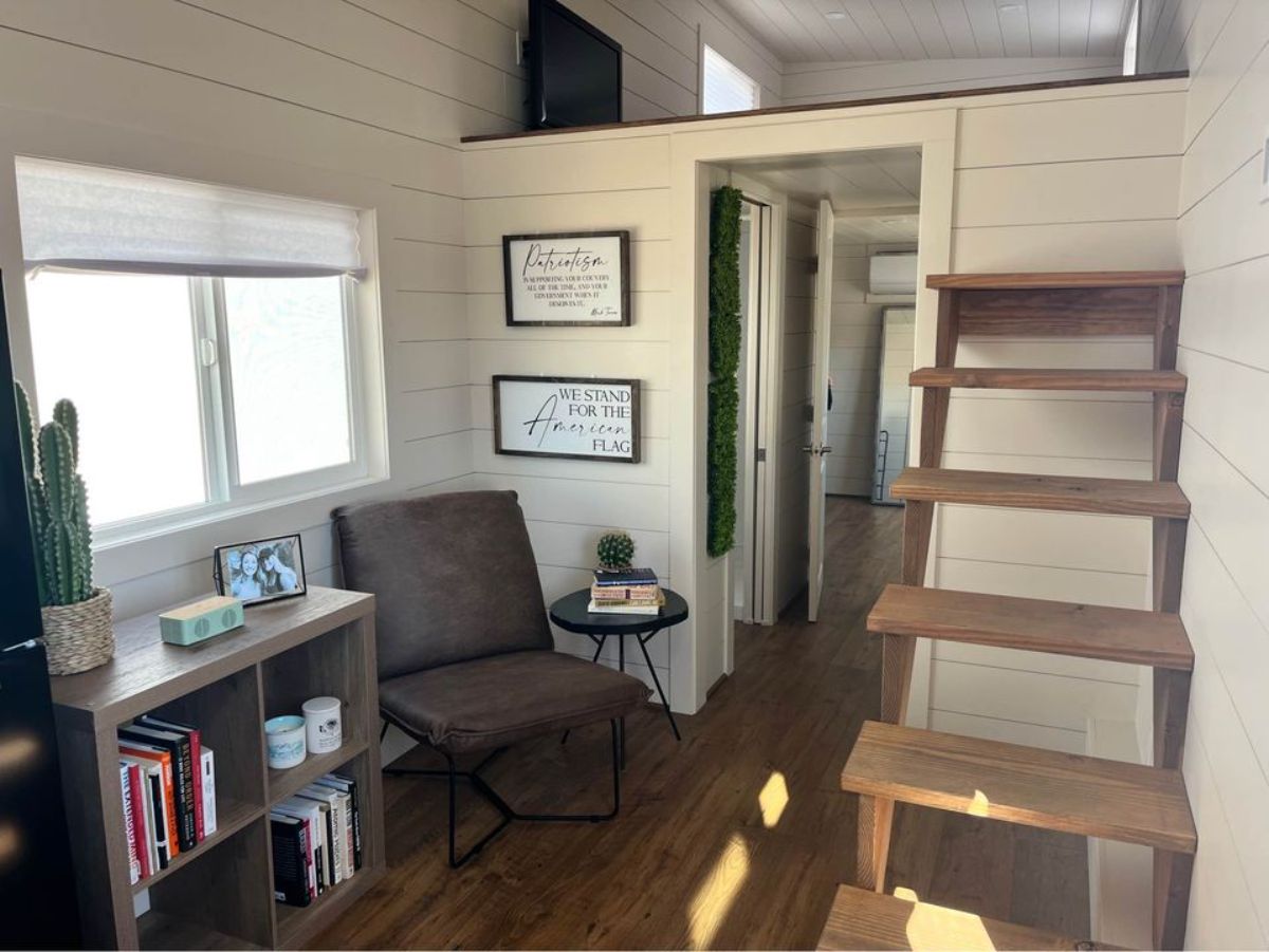 Small and cozy living area of 30’ Modern Tiny House on Wheels has a small chair, a table, book shelf and few wall hanging decorates