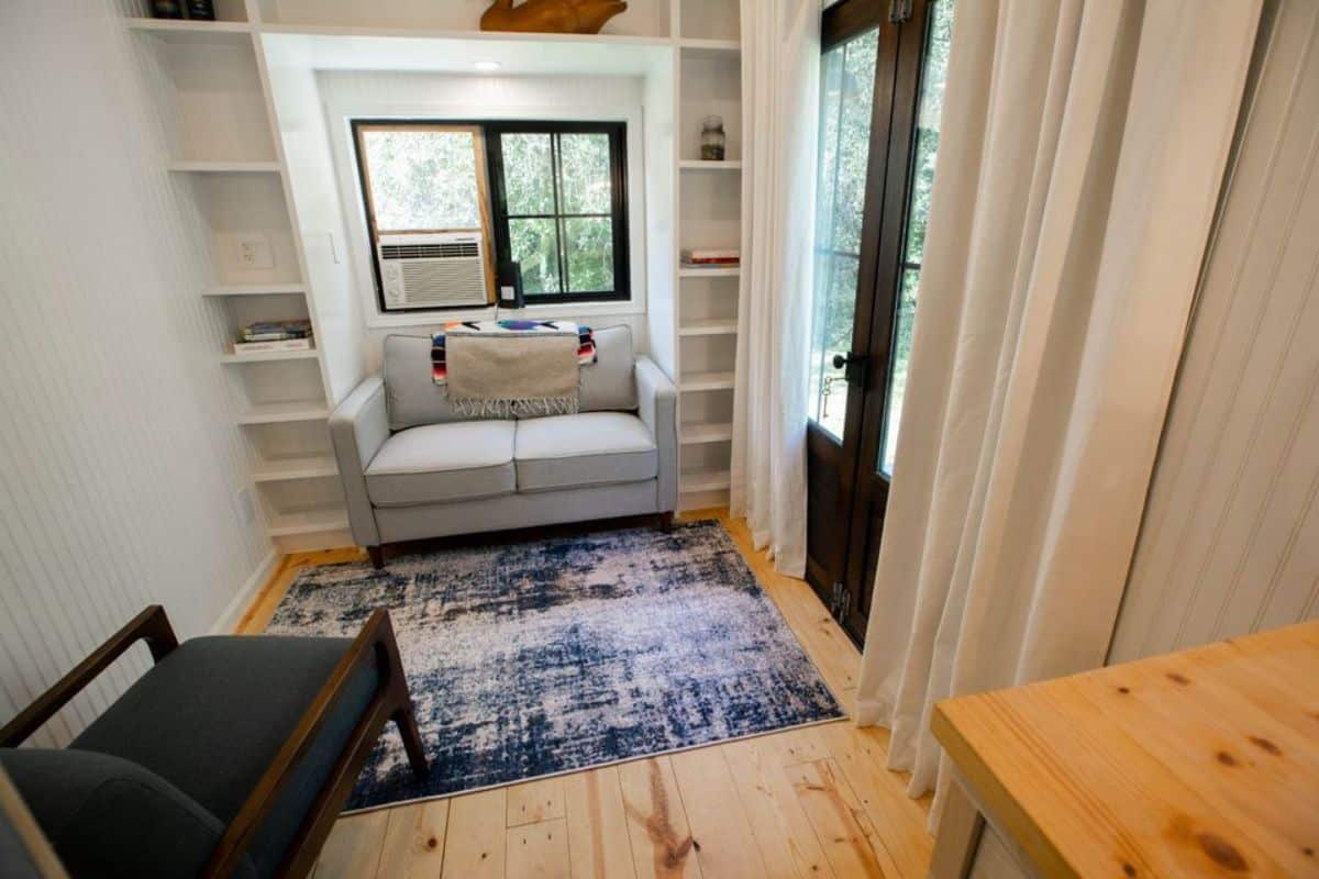 Living area of 20’ Tiny House has a couch, wall mounted book shelf behind and chair