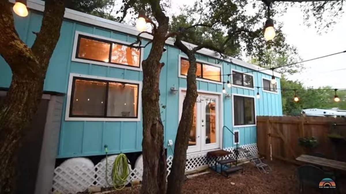 light blue tiny home with lots of windows behind treeline