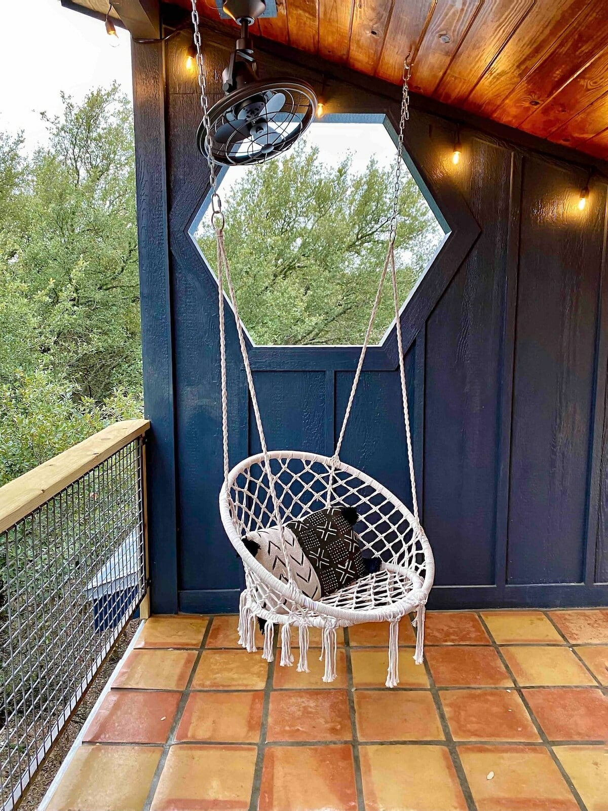 A top covered sleeping porch balcony balcony-side with a hanging round macrame seat, a large circulatory fan mounted on steeply pitched ceilin