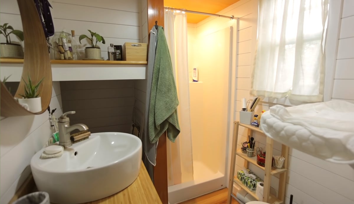 A photo of the bathroom in the tiny house, featuring a shower only, no bathtub, more shelving and storage and a baby changing station over toilet.