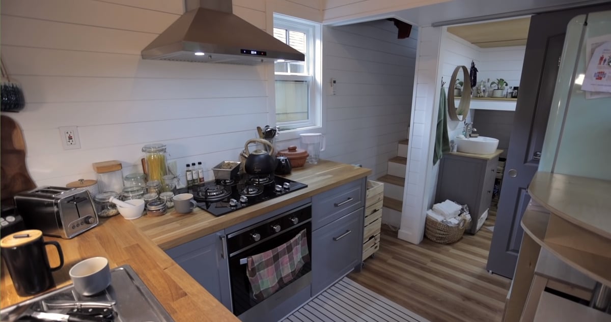 A look at the kitchen of the tiny house, featuring a modern vent hood, gas stovetop, oven and stairway leading to the bathroom