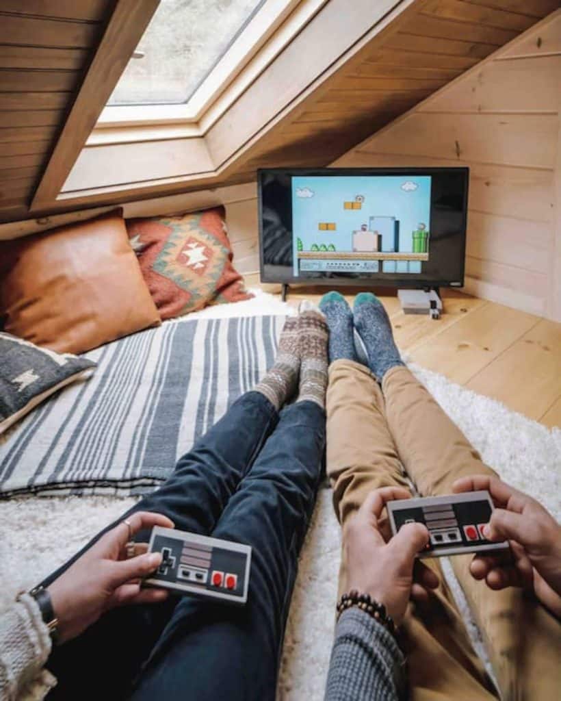 two people holding game controllers on bed in loft with TV in corner of space