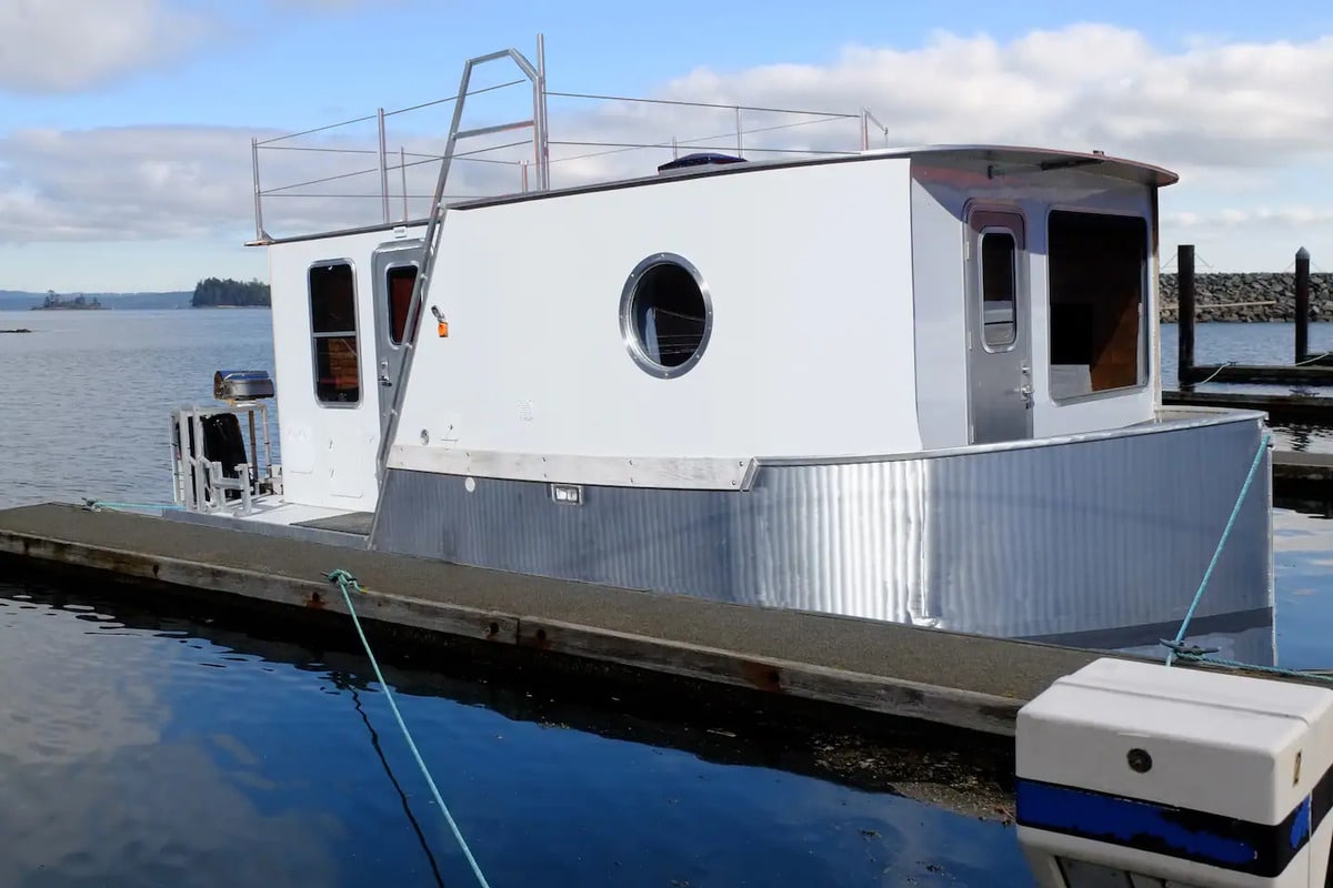 Exterior of a tiny houseboat with round porthole-like windows and an upper deck
