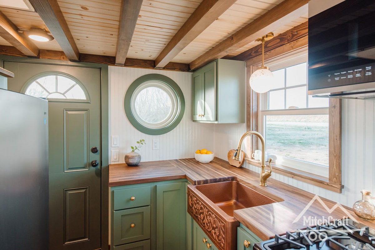 The kitchen at the entryway, featuring an ornate copper sunken farmhouse sink with a modern extendable sprayer faucet, satin finish butcher block countertops all around, a full-sized fridge and a hobbit-y porthole window beside the exterior door over the counter.