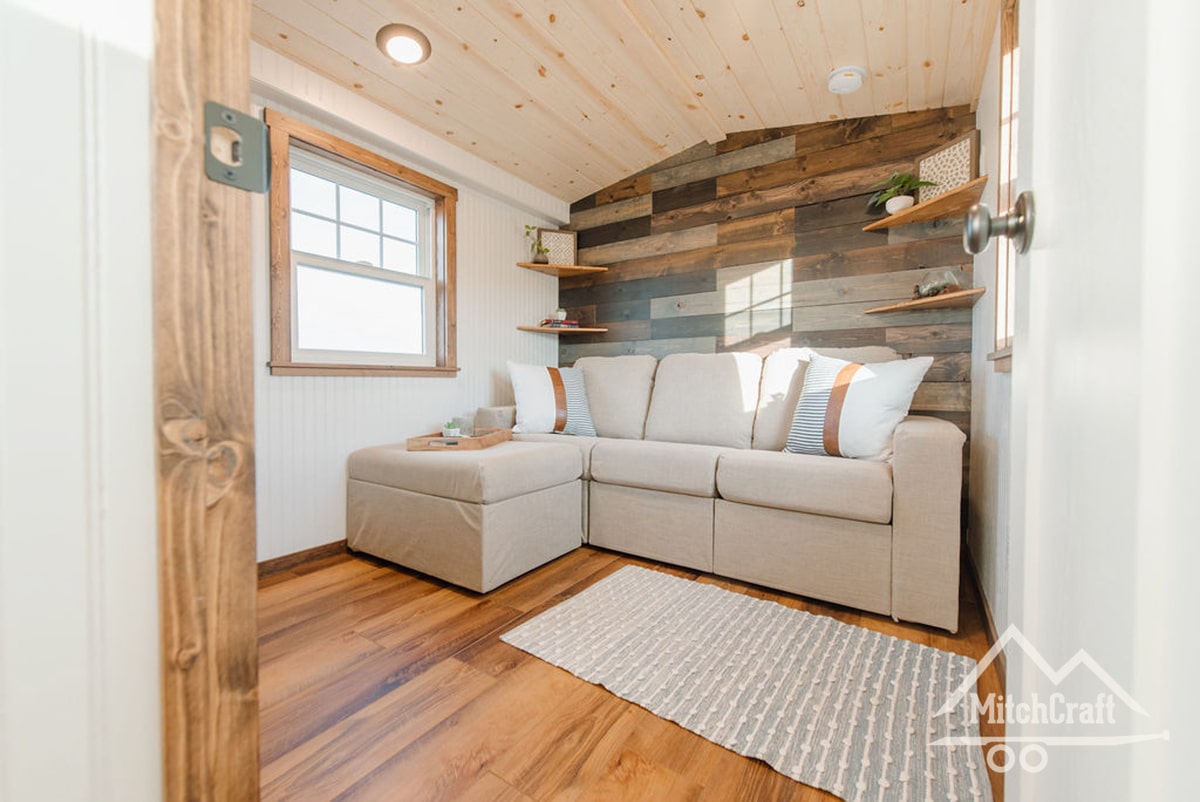 A view of the living area, located at the front of the trailer over the gooseneck, featuring a natural rustic reclaimed wood board mosaic along the back wall, roomy enough for a modest full-sized couch, various stained and sealed, painted, and natural wood tones throughout, and a window for natural light, short LED "can" lights throughout the house in the ceiling