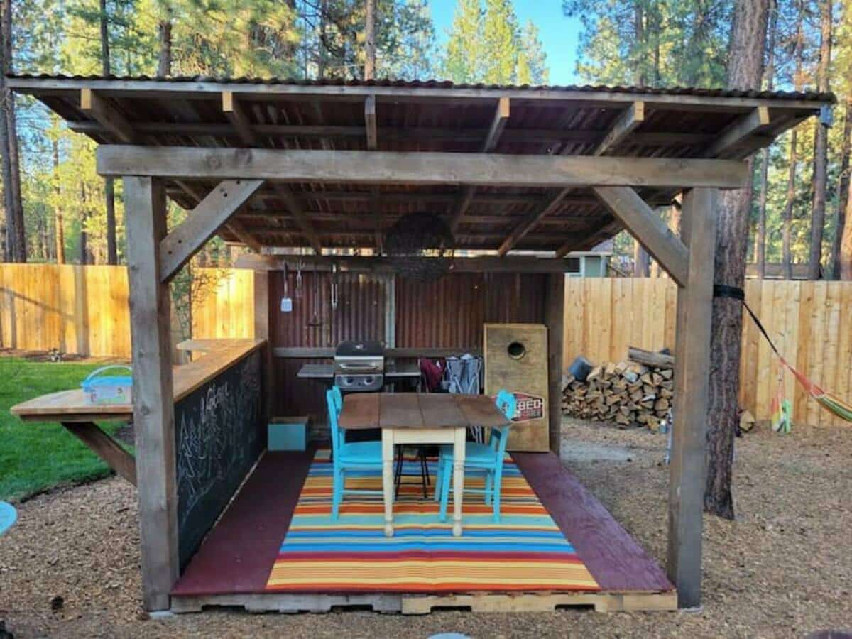 Eclectic covered picnic and bar area with propane grill, bar with chalkboard dividing walk, bean bag toss game, raised wooden deck, and corrugated galvanized steel coverin