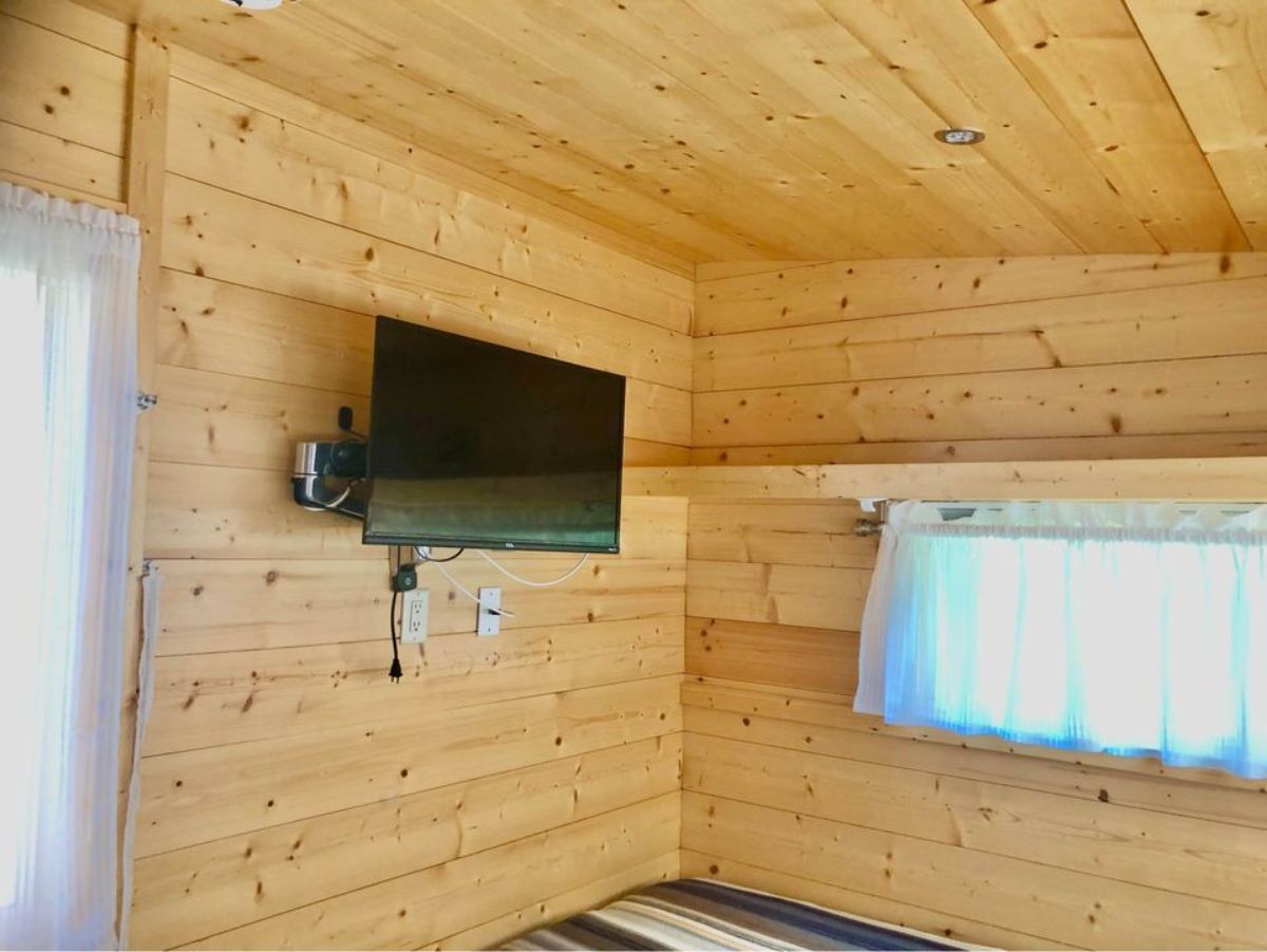 living area of hybrid tiny home has wall mounted TV set opposite to bed